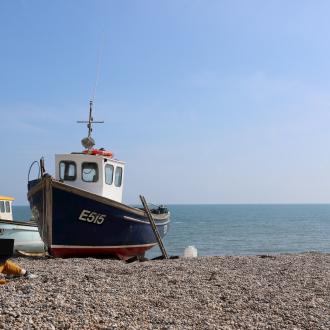 Person sitting on a pebble beach with a laptop next to some moored fishing boats